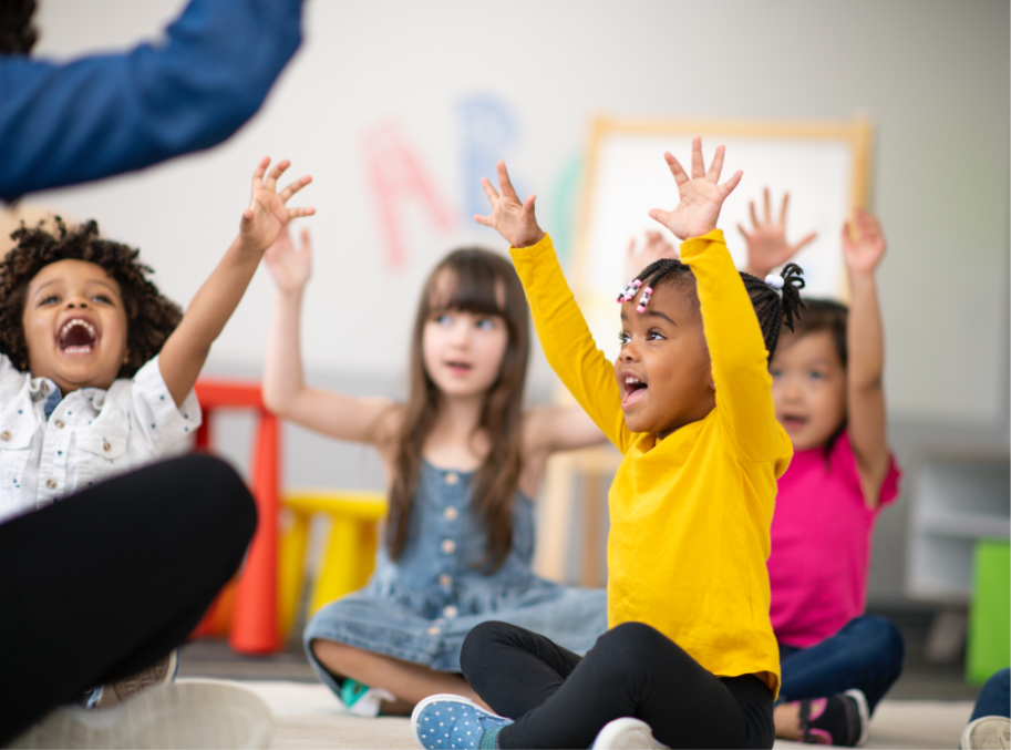 Small children sitting on the floor raising their arms and laughing in school setting