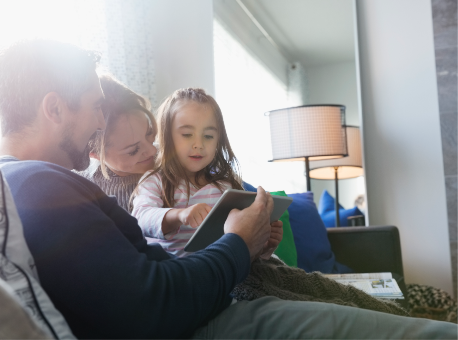 Mom, Dad and young girl sitting on couch looking at tablet and smiling